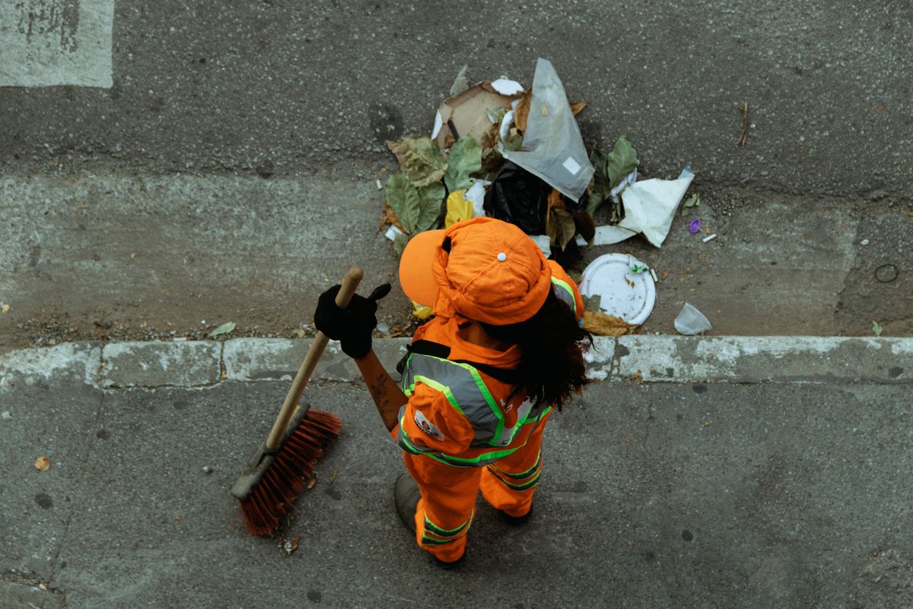 services-03 Top view of a street cleaner wearing orange uniform sweeping debris on a city sidewalk.
