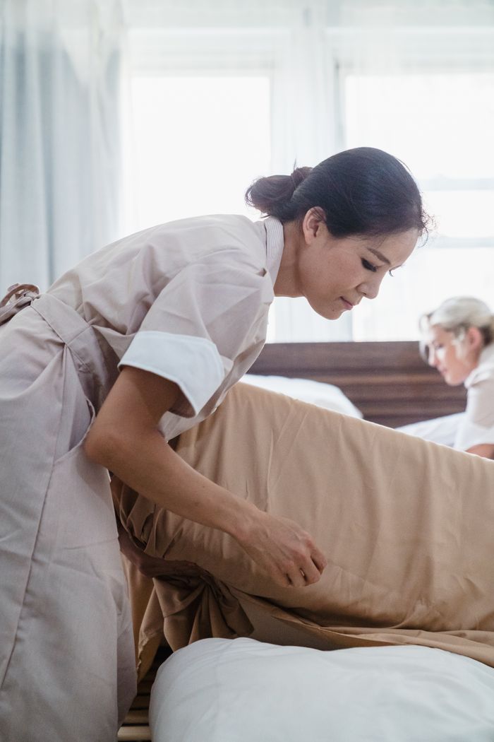 who-we-are Hotel staff in uniform making a bed with precision and care in a bright room setting.